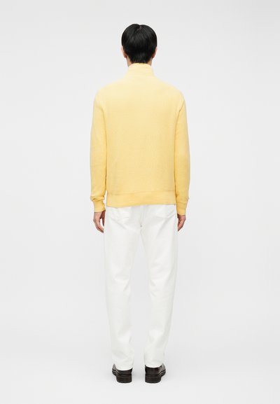 Άνδρας που στέκεται με την πλάτη γυρισμένη φορώντας ένα ανοιχτό κίτρινο πουλόβερ, λευκό παντελόνι και σκούρα καφέ παπούτσια, μπροστά σε έναν απλό φωτεινό φόντο.