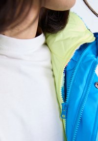 Wendbare Jacke mit einer hellgrünen Außenschicht und einem leuchtend blauen Innenfutter, ausgestattet mit einem Reißverschluss und einer weichen Innenstruktur.