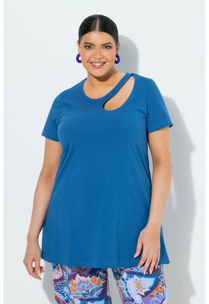 Kobieta o ciemnych włosach, ubrana w niebieską bluzkę z krótkim rękawem i wycięciem na ramieniu oraz wielokolorowe spodnie w kwiatowy wzór, uśmiechająca się na białym tle.