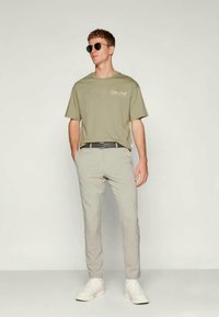 Olivengrønn t-skjorte med tekst, lysegrå bukser, svart belte og hvite joggesko. Modellen har på seg svarte solbriller og står i en nøytral posisjon.