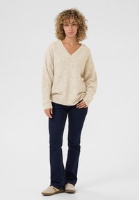 Beige, tekstureret sweater med V-hals og lange ærmer, parret med mørkeblå lige ben jeans og lyse sneakers. Neutral farvepalette.