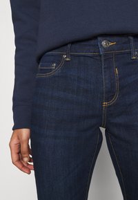 Person wearing dark blue jeans with yellow stitching and a navy blue long-sleeve shirt, hand relaxed by their side.
