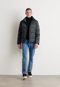 Giacca impermeabile grigia con colletto alto, chiusura con zip, abbinata a jeans blu e scarpe slip-on nere. Materiale texturizzato, design casual.