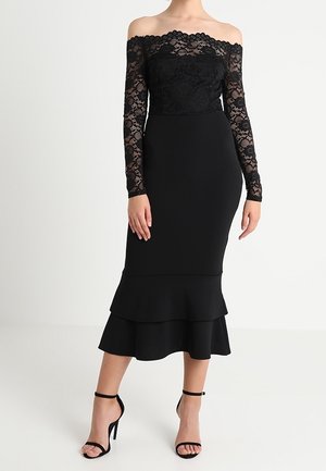 Femme portant une robe noire en dentelle à épaules dénudées avec manches longues et ourlet à volants superposés, associée à des escarpins noirs à bride à la cheville.