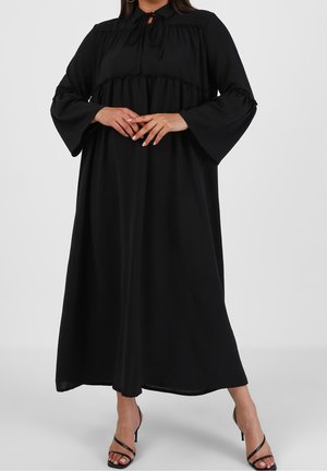 Femme portant une longue robe noire à manches évasées et encolure nouée, assortie à des sandales à talons hauts noires à lanières, sur un fond uni.