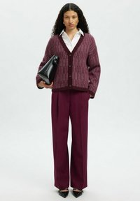 Kobieta w fioletowym swetrze, białej koszuli, szerokich fioletowych spodniach, trzymająca czarną torebkę, stojąca na jednolitym tle.