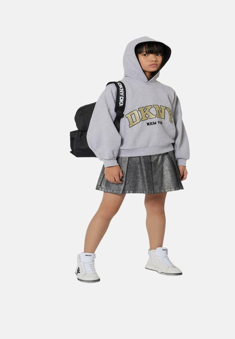Sweat à capuche gris avec "DKNY" en jaune, jupe plissée grise métallisée, baskets blanches et un sac à dos noir avec une bandoulière de marque.