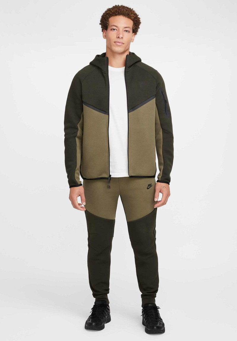 Nike hoodie and sweatpants set in olive green and black. Features a zip-up front, structured design, and logo detail on the pants.