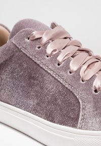 Gros plan sur une chaussure de sport en velours lavande avec des lacets larges en ruban de satin et une semelle en caoutchouc blanche.