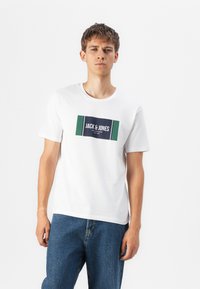 Hvit bomull t-skjorte med et rektangulært grafisk trykk i marineblått og grønt med teksten "JACK & JONES", kombinert med blå denimjeans.