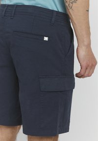 Mørkeblå cargo shorts laget av slitesterk bomullsblanding, med en sidelomme, en baklomme, og sømdetaljer langs sømmene.