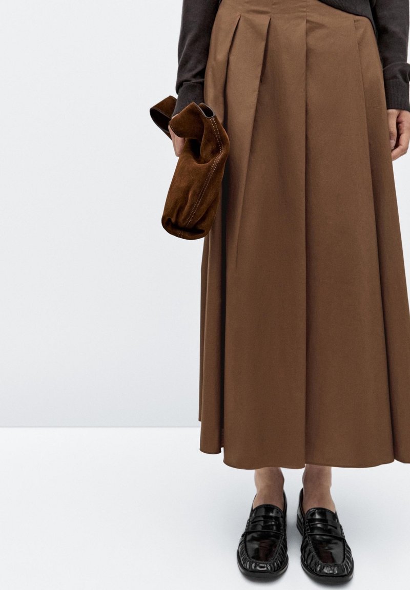 Personne portant une jupe midi plissée marron, des mocassins noirs, et tenant un sac à main en daim marron à côté d'elle.