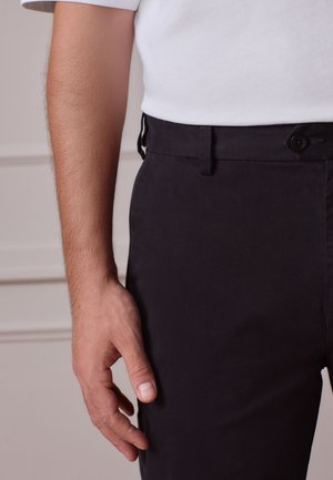 Artimos planas asmens dešinės rankos ir rankos, poilsio padėtyje prie kūno, dėvinčio baltą marškinius ir juodus kelnes su užsegamu juosmeniu.