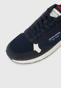 Marineblå sneakers med ruskind og mesh-materialer, rund tå, et hvidt logo og en kontrasterende rød accent på tungen.