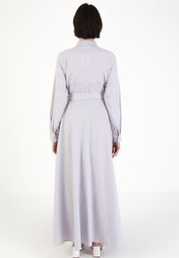 Robe longue gris clair avec un col structuré, des manches boutonnées et une ceinture nouée. Tissu fluide atteignant les chevilles, créant une silhouette élégante.
