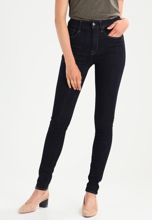 Femme portant un jean skinny foncé avec un haut vert et des chaussures beiges en daim à talons blocs, une main dans la poche et l'autre reposant sur la cuisse.