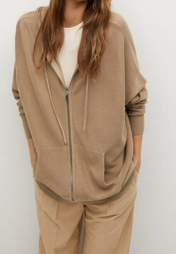 Femme portant un sweat à capuche beige zippé avec poches avant et cordons, assorti à un pantalon beige ample, debout devant un fond uni.