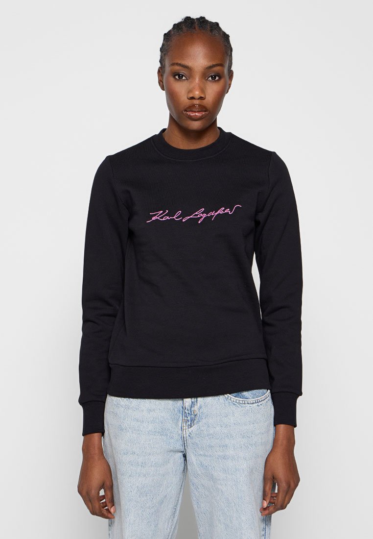 Schwarzer Sweatshirt mit gerippter Textur, versehen mit pink gesticktem "Karl Lagerfeld" Logo über der Brust. Kombiniert mit hellblauen Jeans.