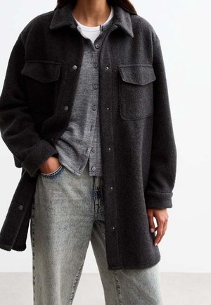 Femme portant des vêtements gris superposés comprenant un manteau en laine gris foncé, un cardigan boutonné, un haut blanc et un jean bleu délavé.
