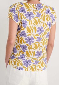 Donna che indossa una maglietta a maniche corte con un motivo floreale giallo e viola, in piedi con le mani nelle tasche dei pantaloni bianchi, di spalle rispetto alla fotocamera.