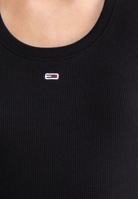 Nærbillede af sort ribbet stoftop med lille rektangulært rødt, hvidt og navy logo syet under en rund halsudskæring.