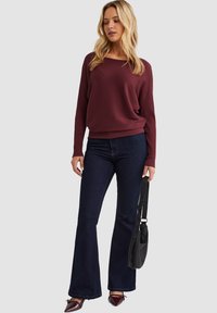 Bordeaux ribbeltrui, relaxte pasvorm, gecombineerd met donkere flare jeans en puntige hakken. Een zwarte schoudertas maakt de outfit compleet.