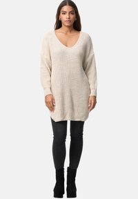 Suéter de punto beige con un escote en V profundo, corte oversize, patrón texturizado y puños de canalé, combinado con jeans ajustados negros y botas.
