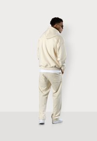 Sweat à capuche beige avec une coupe décontractée, associé à un jogging assorti avec de grandes poches arrière, porté avec des sneakers blanches.