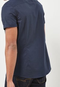 Camisa de manga corta en color navy, hecha de algodón con una textura suave. Presenta un cuello clásico y un dobladillo curvado. La vista trasera muestra una costura cuidada.
