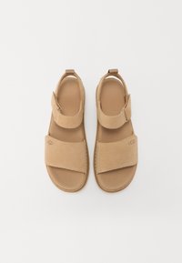 Šviesiai rudos suknelės šlepetės su reguliuojamomis sagtimis, plokščiu padu ir tekstūruotu padu. UGG logotipas išspaudžiamas šone. Minimalistinis dizainas.