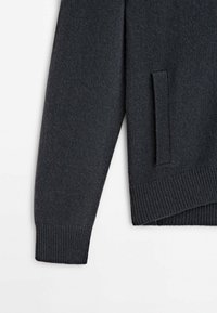 Manche et poche latérale d’un pull en maille gris charbon avec des poignets et un ourlet côtelés sur fond blanc.