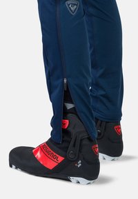 Laivastonsiniset pehmeät kuorihousut, joissa on vetoketjuja, ja mustat hiihtokengät, joissa on punaisia yksityiskohtia ja teksturoitu pinta, esittäen brändäysdetaljeja.