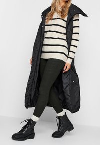 Gilet nero imbottito lungo, maglione oversize a righe crema, leggings a costine verde scuro e stivali da combattimento neri con lacci e calzini bianchi.