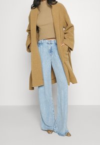 Cappotto oversize color cammello realizzato in tessuto morbido, abbinato a un crop top beige a coste e jeans a gamba larga azzurri. Texture evidente e vestibilità comoda.