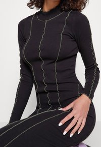 Schwarzes, geripptes Langarmkleid mit grünen, wellenförmigen Steppdetails. Stehkragen mit gewellten Kanten und taillierte Silhouette.