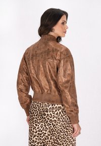 Jachetă bomber din piele maro, cu guler și manșete ribbate, având o textură ușor uzată. Purtați peste o fustă cu imprimeu leopard.