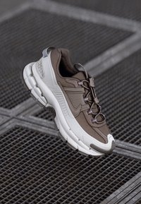 Brūni un balti sporta apavi ar sietu un sintētisku augšu, biezs zoles un aukliņu dizains. Iespējas ar strukturētām rakstu un šuvju detaļām.