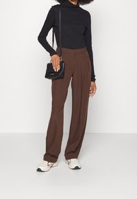 Braune, hoch taillierte Hosen mit Falten, kombiniert mit einem schwarzen langärmligen Rollkragenpullover und weißen Turnschuhen. Eine kleine schwarze Umhängetasche wird gehalten.