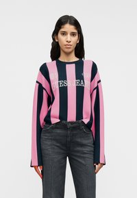 Ριγέ πουλόβερ πλεκτό σε ροζ και ναυτικό μπλε με στρογγυλή λαιμόκοψη, μακριά μανίκια, εκτύπωση λογότυπου και ρι μπ μανσέτες. Συνδυασμένο με σκούρο γκρι τζιν από denim.