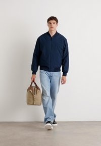 Veste en cuir d'aviateur navy avec une fermeture éclair frontale, des poignets et une taille côtelés ; jeans bleu clair ; sac beige avec des accents marron ; baskets blanches.