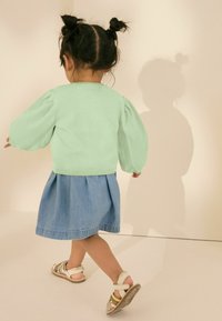 Mintgroene gebreide cardigan met pofmouwen, gedragen met een denimrok. Het kind draagt meerkleurige sandalen met bandjes.