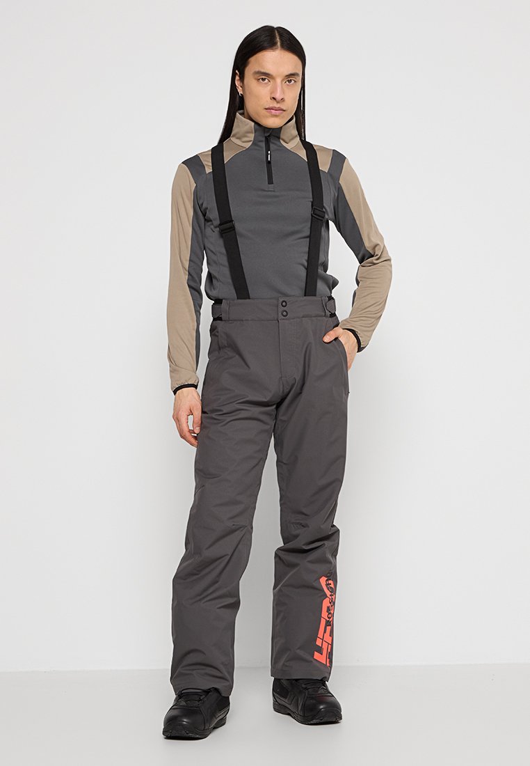 Szare spodnie narciarskie ze szelkami, zapięcie na guzik i pomarańczowy detal z logo. Noszone z szaro-beżową bluzą z długim rękawem z pół zamkiem.