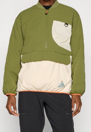 Uomo che indossa un pullover trapuntato verde e beige con mezza zip, tasca sul petto con asola e pantaloni neri con tasche con zip.