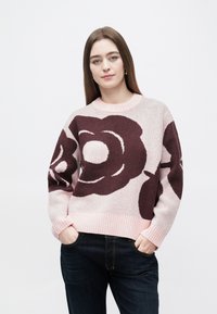 Pleteni pulover v svetlo roza barvi z velikim temno bordo vzorcem cvetja. Manšete in spodnji del z rebrastim vzorcem, sproščen kroj, mehka tekstura. Pod puloverjem temne kavbojke.