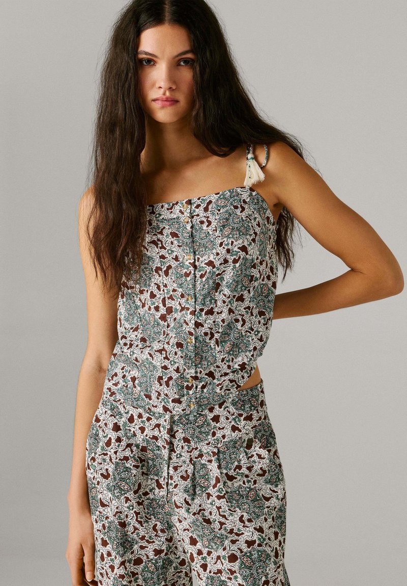 Noor naine, kellel on pikad tumedad juuksed, kannab varrukateta lillemustriga toppi ja sobivaid kõrge vöökohaga pükse, seisab ühe käega puusal.