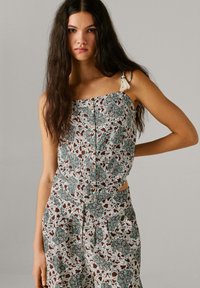 Jonge vrouw met lang donker haar, gekleed in een mouwloze top met bloemmotief en bijpassende hoge taille broek, staand met één hand op haar heup.