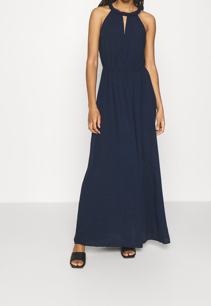 Femme portant une robe longue bleu marine dos nu avec un décolleté en forme de trou de serrure et des sandales noires à talon bloc et bout ouvert.