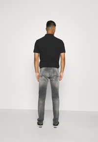 Hombre vistiendo una camisa polo negra, jeans gris desgastados y zapatillas negras, de pie frente a una pared blanca lisa.