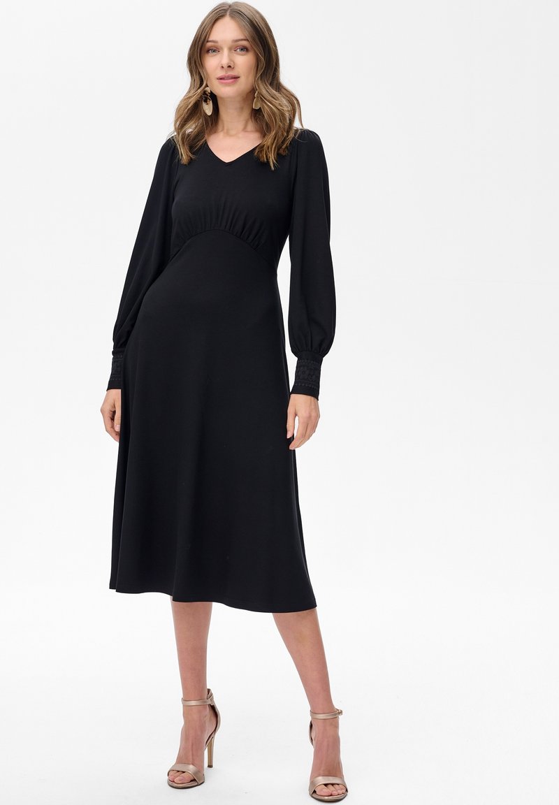 Kobieta w długiej czarnej sukience midi z dopasowaną talią i detalami przy mankietach, w połączeniu z beżowymi sandałami na wysokim obcasie, stojąca na białym tle.