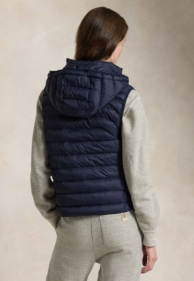 Vrouw met bruin haar die een marineblauw doorgestikt hooded vest draagt over een lichtgrijze sweatshirt en bijpassende joggingbroek, met haar rug naar de camera gericht.